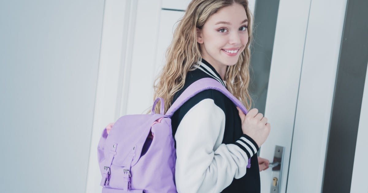 Estudante de pé sorrindo, usando uma mochila lilás, com uma mão segurando uma alça da mochila e a outra na maçaneta da porta.