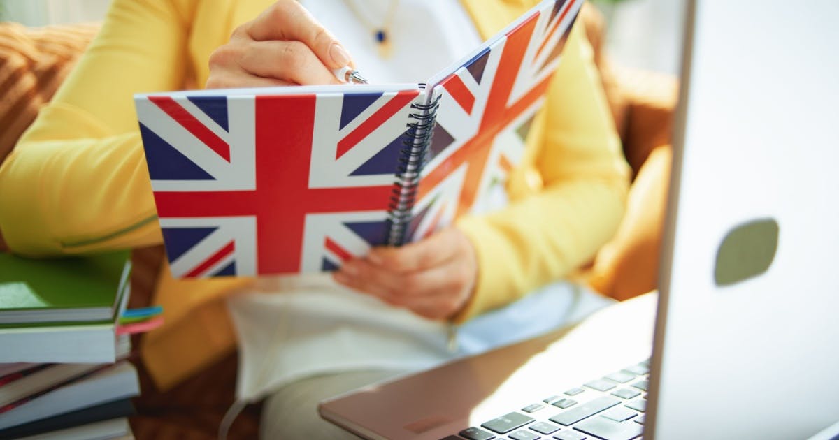 Pessoa estudando sobre como abrir uma escola, na frente de um notebook e segurando um caderno com a bandeira da Grã-Bretanha.