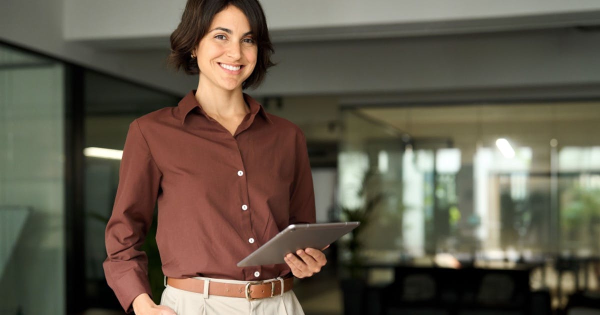 Franqueada de camisa bordô, de pé em uma empresa, sorrindo, enquanto segura um tablet.