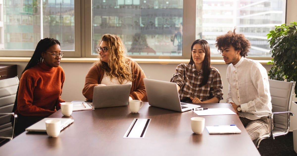 Três mulheres e um homem em uma sala empresarial, sentados diante de uma mesa de reuniões, analisando sobre como empreender.