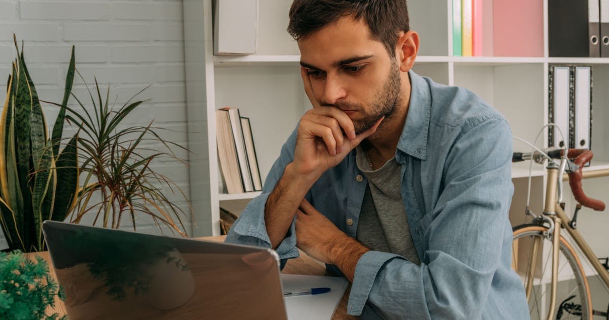 Homem sentado de frente para um notebook, estudando sobre como empreender, com uma prateleira com livros atrás.