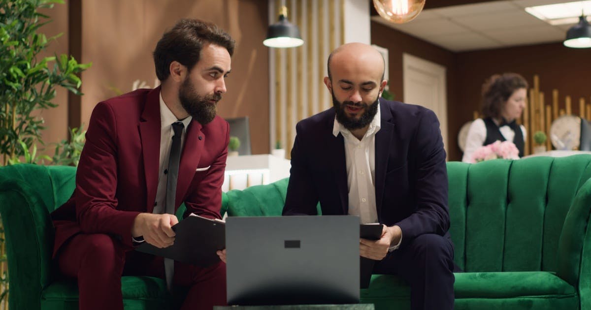 Dois homens de terno sentados em um sofá verde diante de um notebook, conversando sobre como empreender.