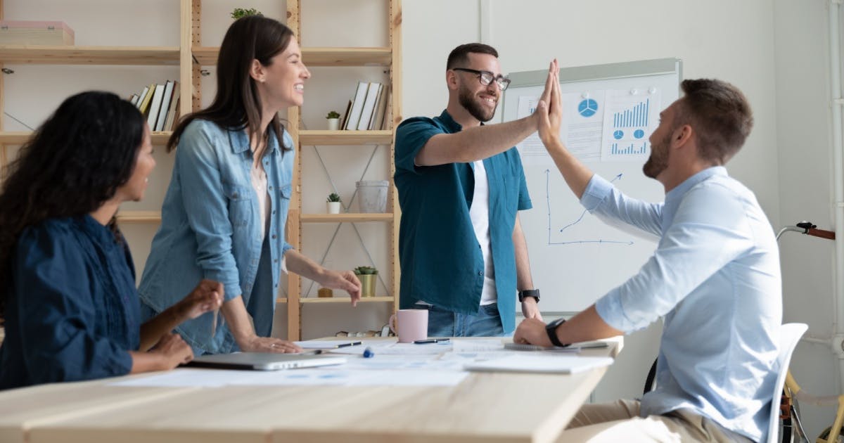 Várias pessoas ao redor de uma mesa, uma pessoa cumprimentando outra com um high five.
