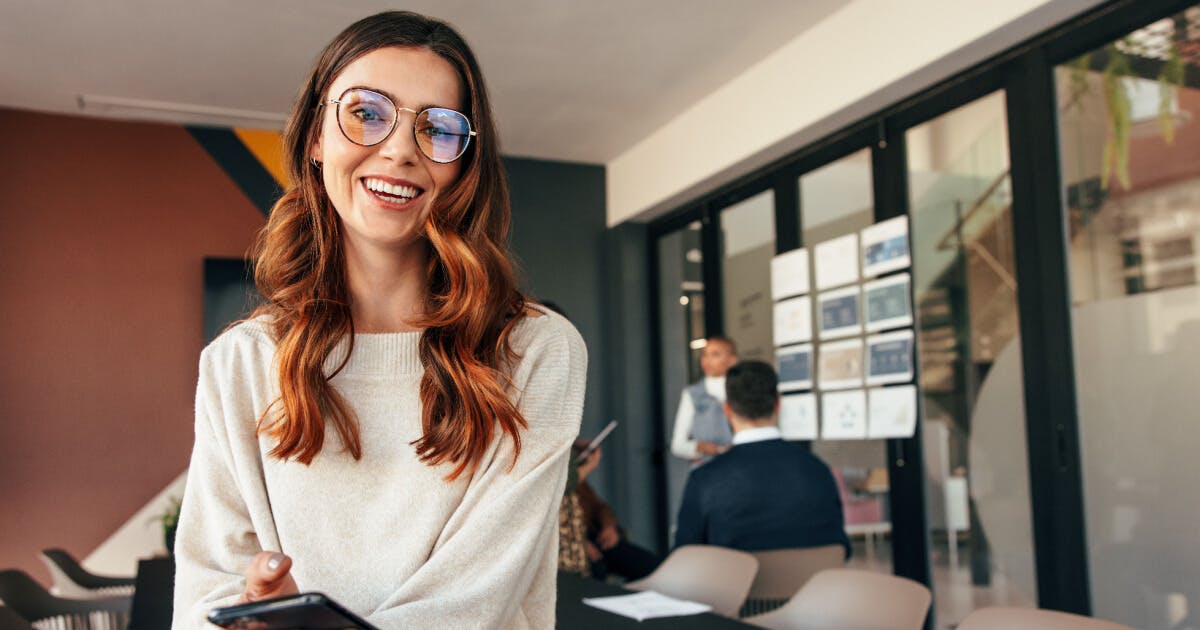 Uma mulher de óculos está em pé em um escritório, com um ambiente profissional ao fundo.