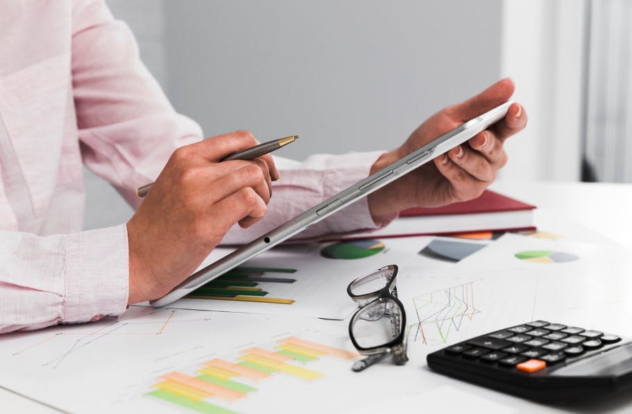Imagem de pessoa usando um tablet para análise de dados e gráficos, com óculos, calculadora, caneta e documentos na mesa de escritório, ideal para negócios e análise financeira.
