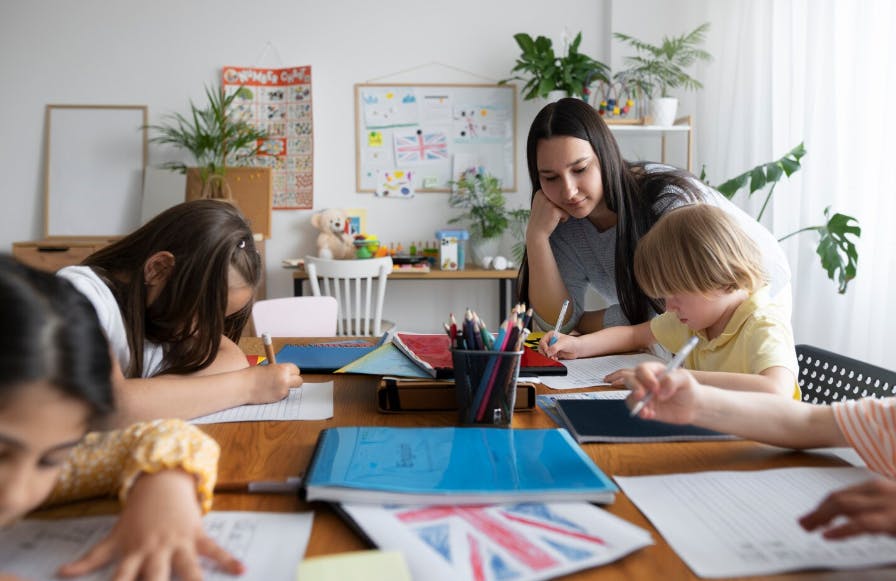 Professora ajudando crianças durante aula em sala de aula, com materiais escolares e quadro de atividades ao fundo.