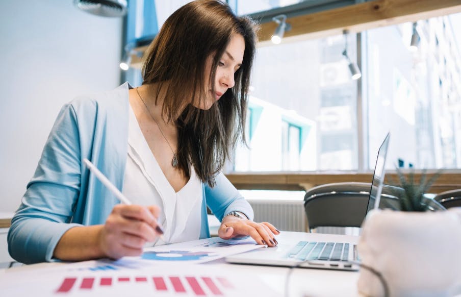 Mulher de cabelos castanhos escuros trabalhando em um laptop e utilizando uma caneta em uma mesa de escritório com ambiente bem iluminado e moderno