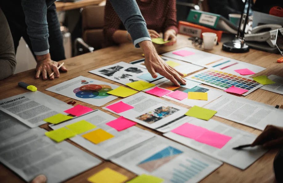 Equipe de profissionais analisando e organizando documentos, gráficos e post-its coloridos em uma reunião de planejamento estratégico.
