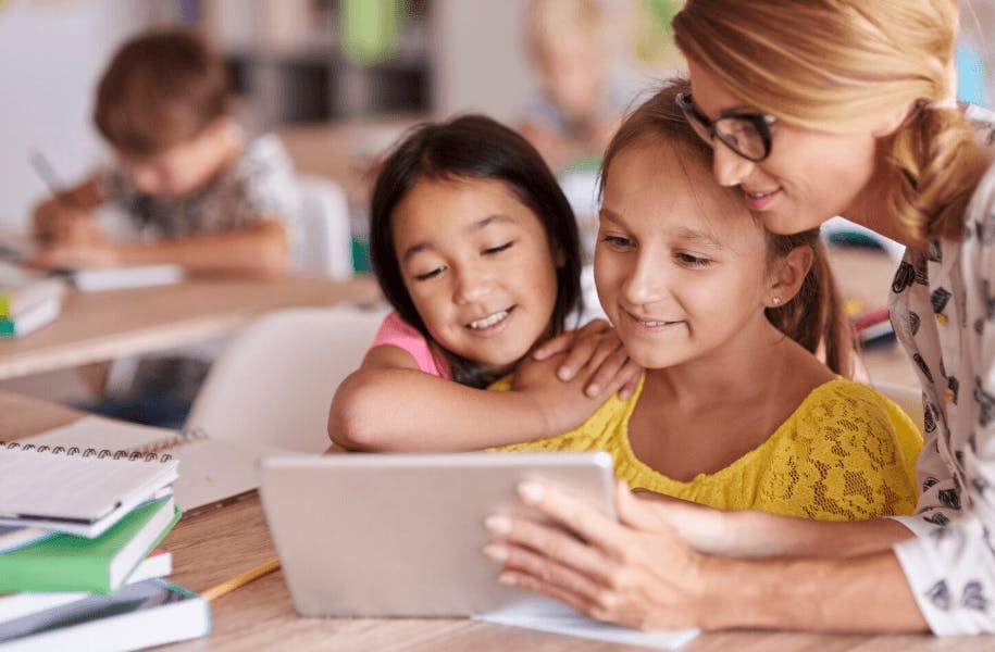 Duas crianças e uma professora observando uma tela de tablet em uma sala de aula, promovendo educação infantil de qualidade para crianças.