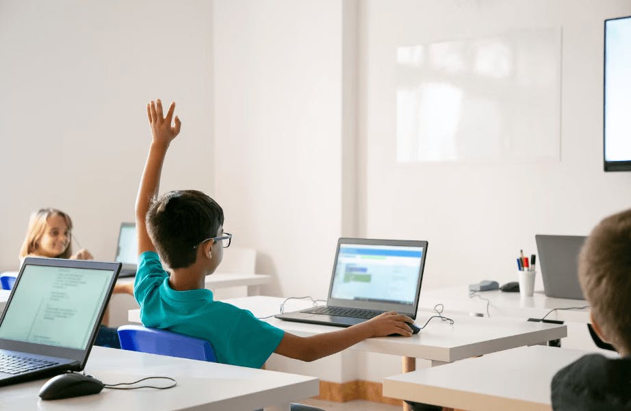 Crianças participando de aula em sala de aula moderna com computadores, professor na frente e alunos atentos. Ambiente de educação inovadora.