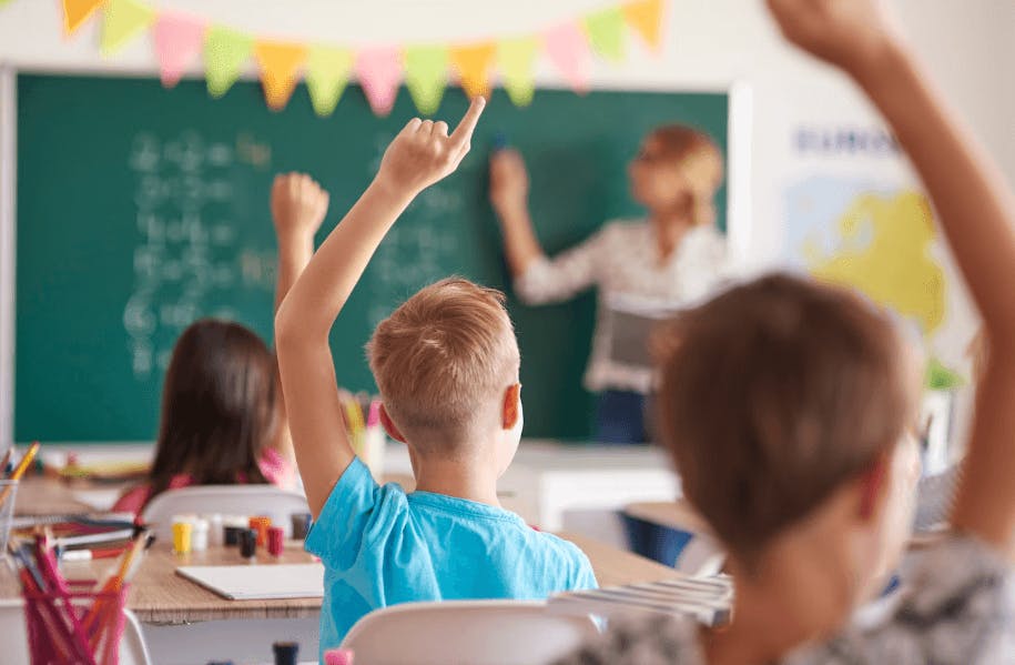 Criança levantando a mão na sala de aula enquanto a professora ensina na lousa ao fundo, ambiente escolar com estudantes participando ativamente.