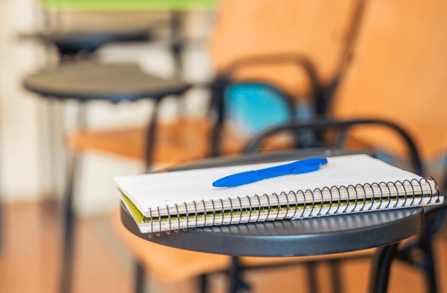 Caderno com uma caneta azul sobre um pencil case de uma sala de aula, com cadeiras ao fundo.