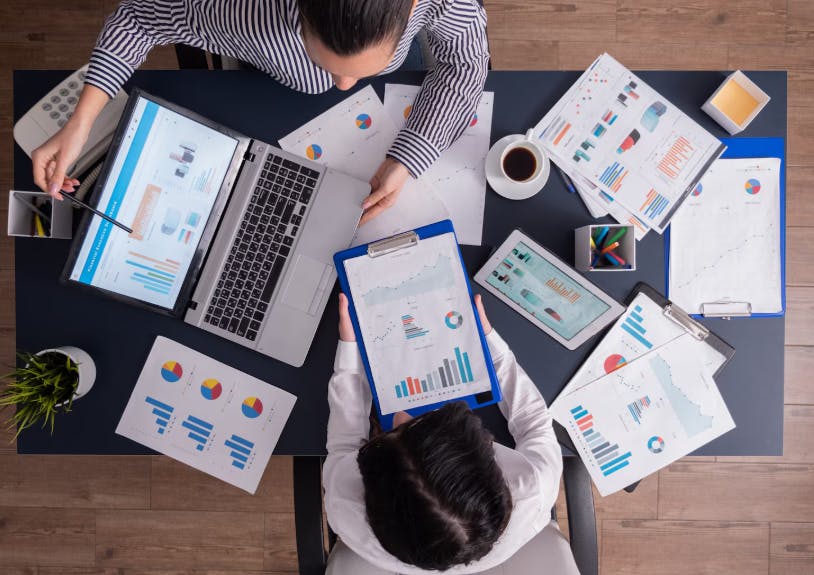 Análise de dados financeiros e gráficos em uma reunião de negócios, com pessoas utilizando laptops, tablets e papéis preenchidos com gráficos e relatórios.