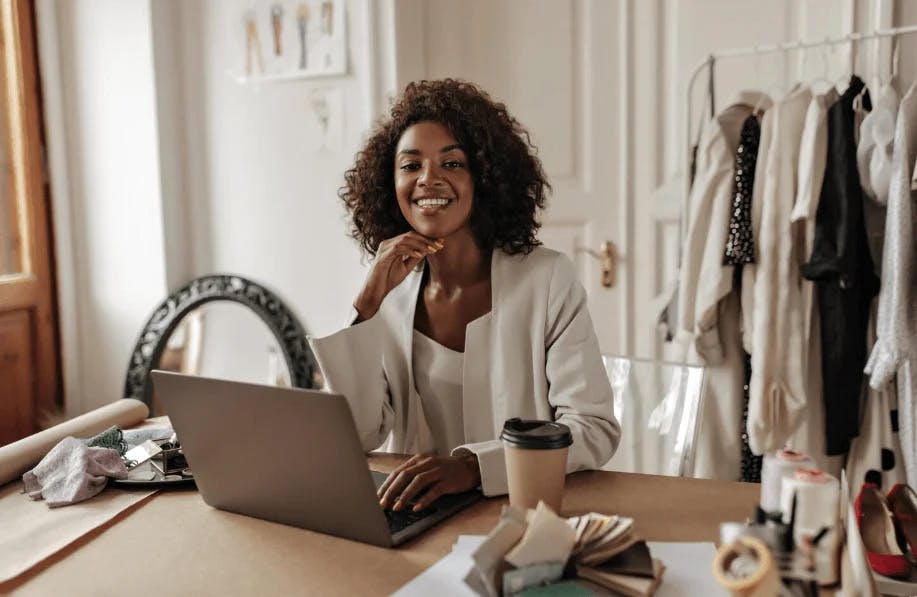 Mulher negra sorridente trabalhando em um escritório de moda com roupas penduradas ao fundo e uma mesa com diversas ferramentas de costura e acabamento.