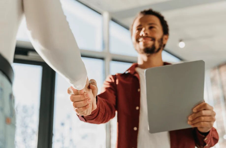 Homem de negócios sorridente apertando a mão de outra pessoa durante uma reunião em um ambiente moderno com janelas grandes ao fundo, simbolizando sucesso e parcerias.