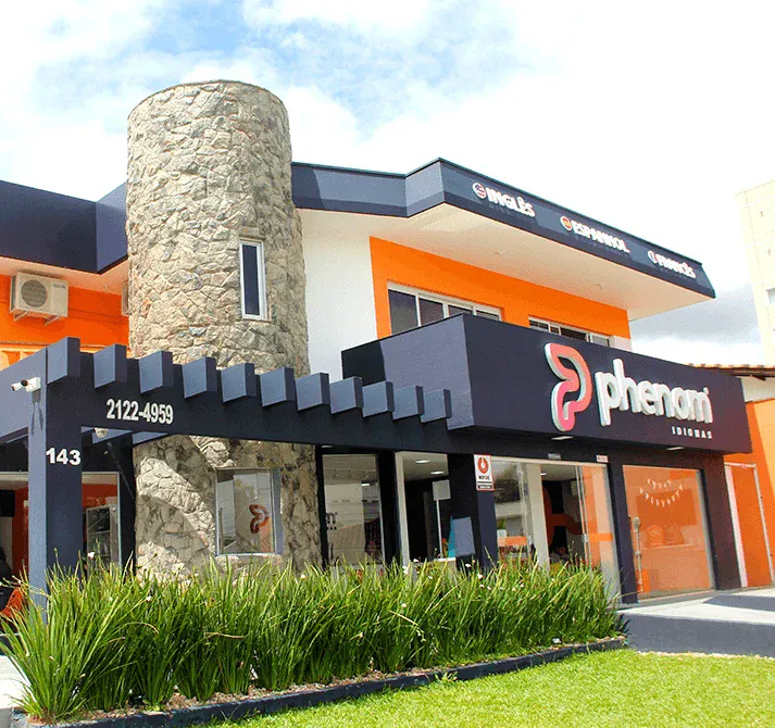 Fachada de uma loja Phenom com letreiro em destaque, parede de pedra e prédio comercial moderno em ambiente urbano, com área externa e gramado em frente.