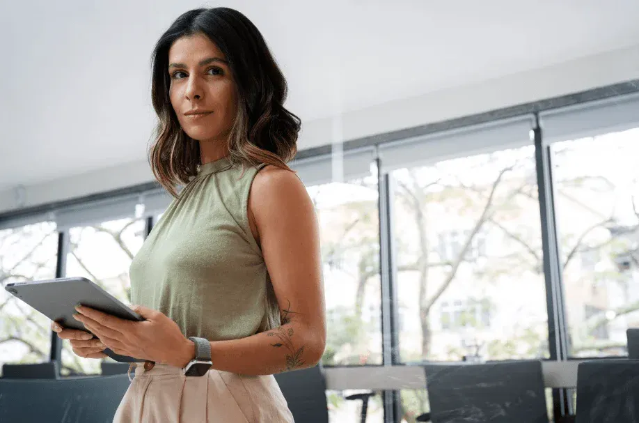Mulher em ambiente corporativo, de pé ao lado de janelas, segurando uma prancheta e olhando para o lado com expressão confiante. Usa vestido verde claro e relógio no pulso.