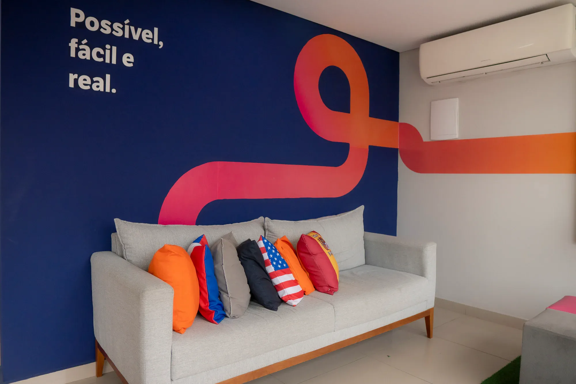 Sala moderna decorada com sofá cinza e almofadas coloridas, parede azul com grande letra em destaque e frase “Possível, fácil e real”, além de ar-condicionado e piso claro.
