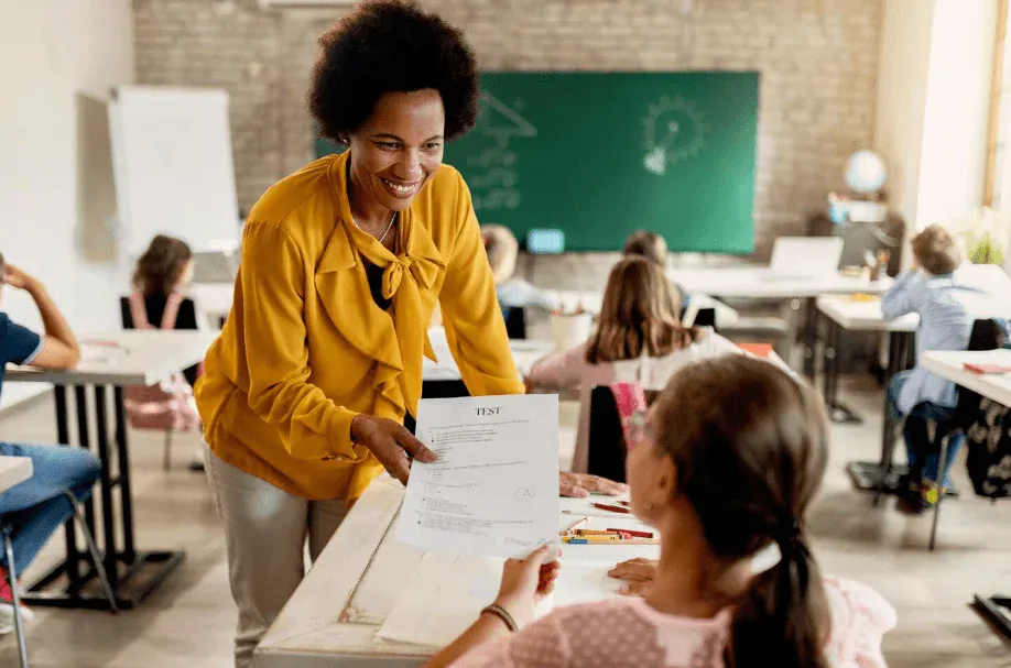 Professora em sala de aula sorrindo enquanto entrega uma prova impressa para alunos sentados em mesas, em um ambiente de ensino com quadro verde ao fundo.