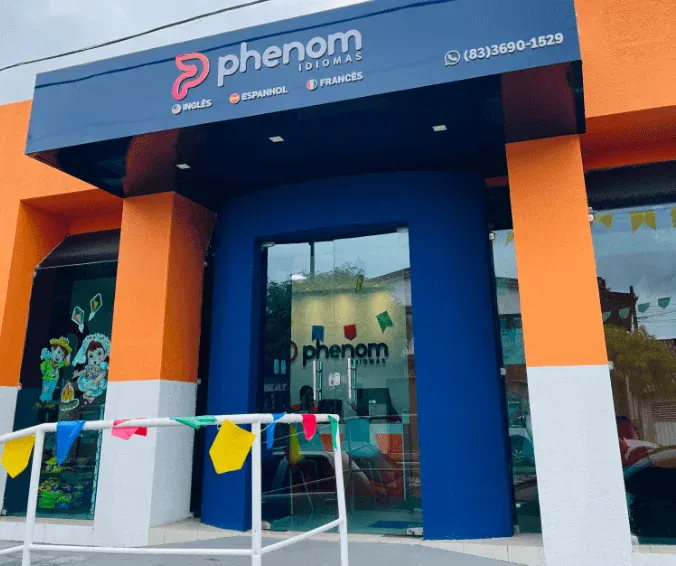 Fachada da loja Phenom com letreiro azul e logo rosa, entrada com colunas laranja e vidro refletivo, além de bandeirinhas coloridas na frente do prédio.