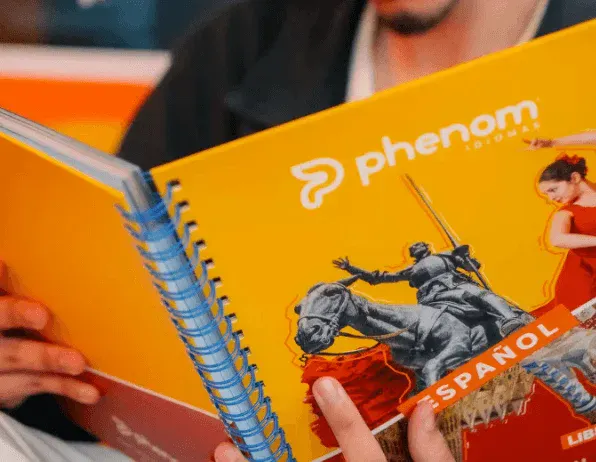 Uma pessoa segura e lê um livro amarelo de capa espiral para aprendizado da língua espanhola intitulado "fenômeno", que apresenta imagens de uma estátua e uma dançarina na capa