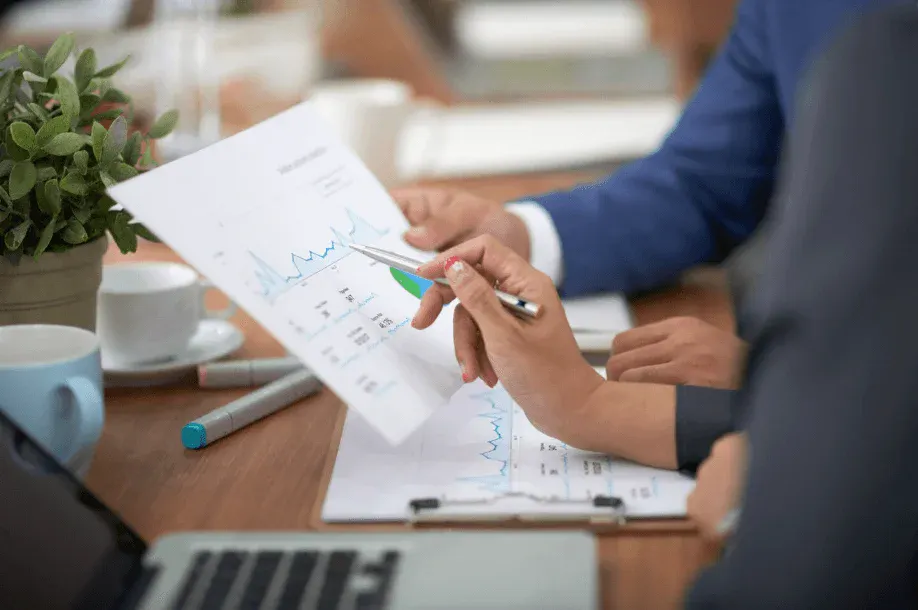 Pessoa analisando um gráfico impresso em reunião de negócios, com caneta apontando dados e outras folhas na mesa, enquanto um parceiro acompanha o material. Ambiente corporativo.