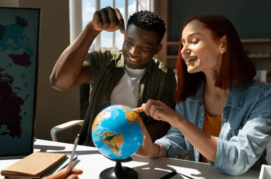 Jovens conversando em uma sala de estudos, enquanto um deles levanta o punho e a outra aponta para um globo terrestre sobre a mesa, sugerindo aprendizado e trabalho em equipe.