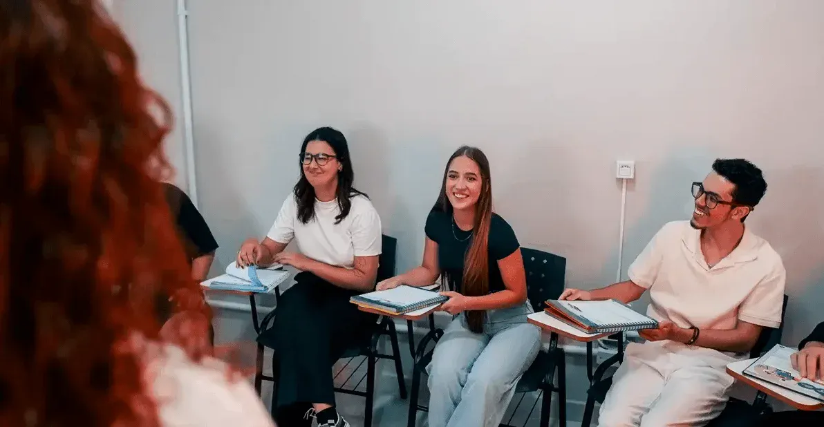 Reunião em sala de aula com quatro pessoas sentadas ao redor de mesas, conversando e estudando com cadernos e anotações, em ambiente interno com parede clara.