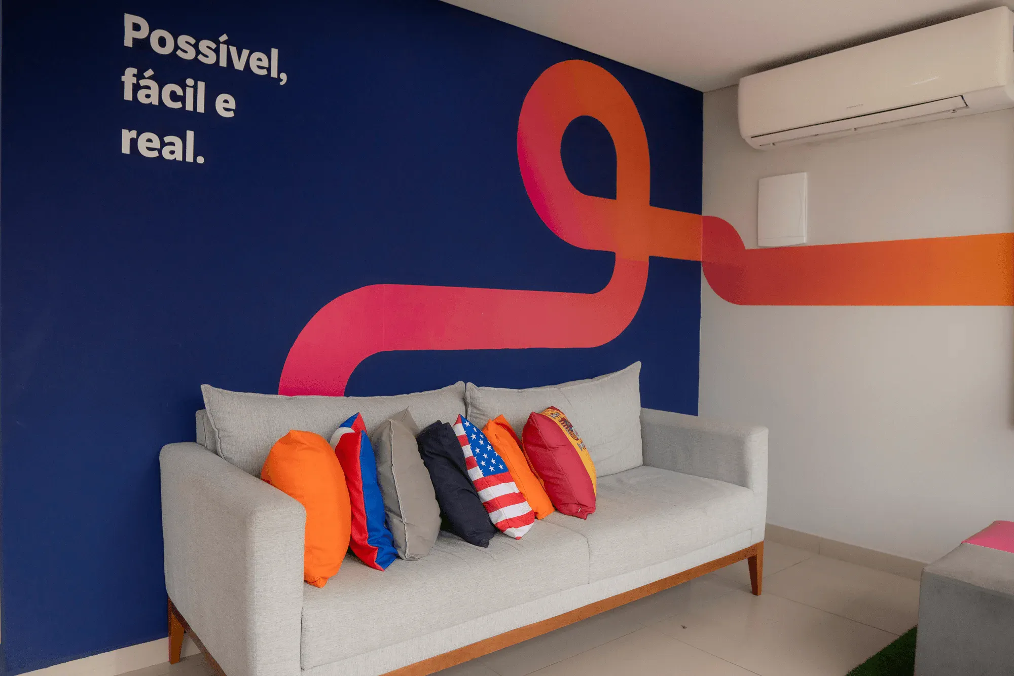 Sala de estar com sofá cinza, almofadas coloridas e parede azul com lettering “Possível, fácil e real.”, além de símbolo estilizado em vermelho e faixa laranja. Há ar-condicionado branco na parede.