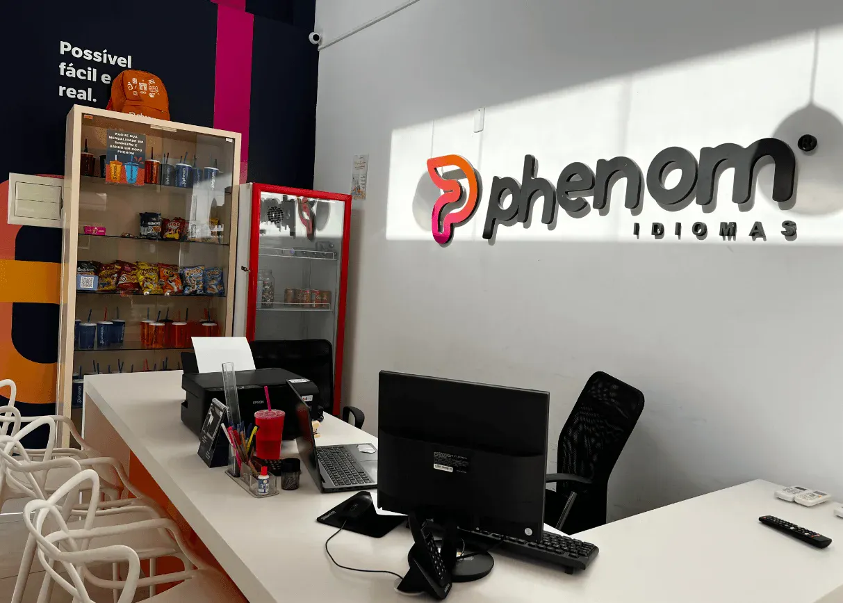 Ambiente de atendimento da loja Phenom com logo na parede, mesa de escritório com computadores, cadeiras e armários com produtos e bebidas ao fundo.