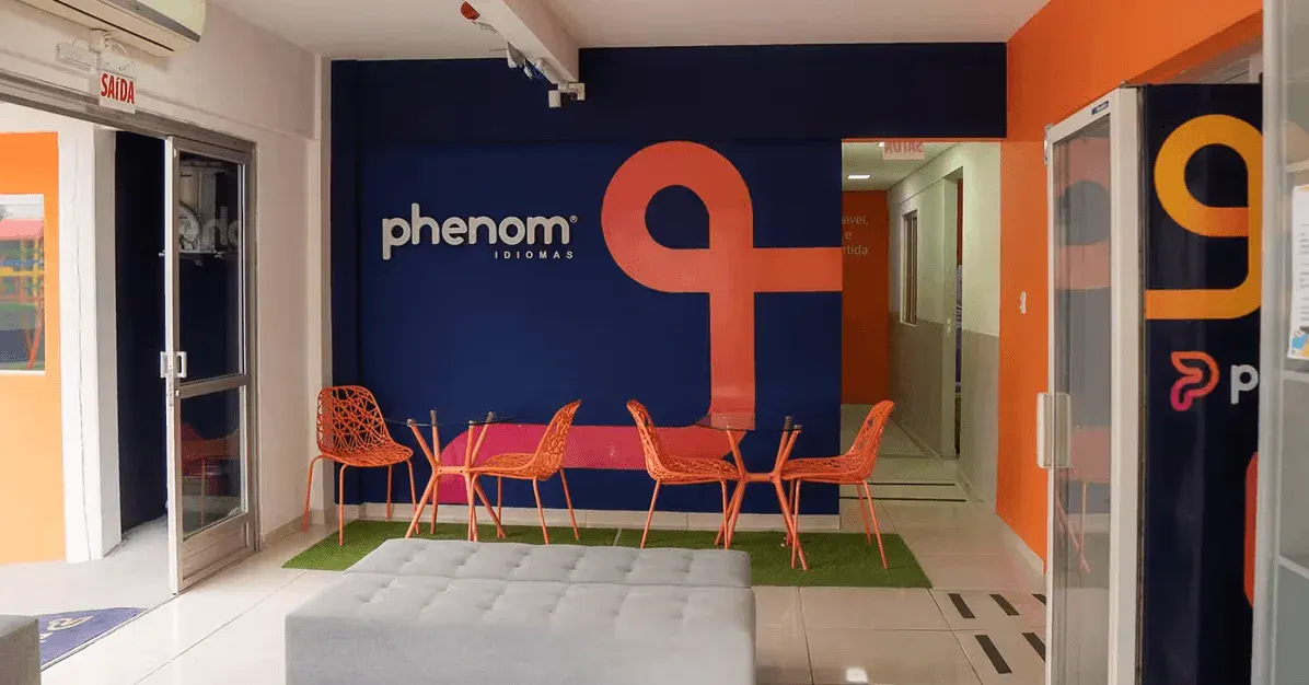 Corredor de escritório com paredes laranja e azul-marinho, logo “phenom” e número 9, cadeiras laranja e mesa ao fundo, piso revestido e tapete verde com iluminação embutida no teto.
