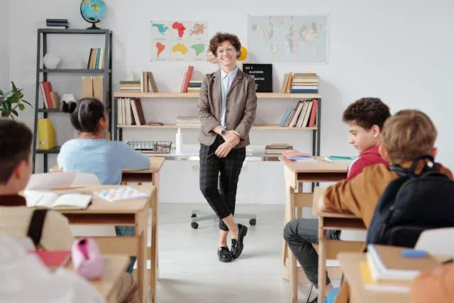 Professora em sala de aula com alunos sentados em carteiras, em um ambiente escolar organizado com mapas e estantes ao fundo, durante aula e orientação.