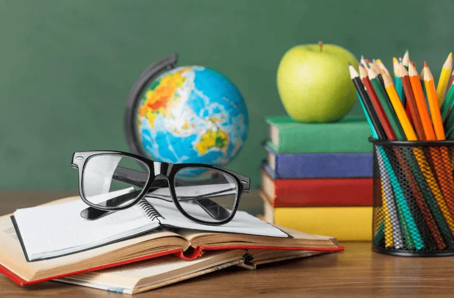 Cena de material escolar sobre uma mesa: óculos, livros abertos, lápis coloridos em um estojo e globo terrestre ao lado, com maçã amarela ao fundo.