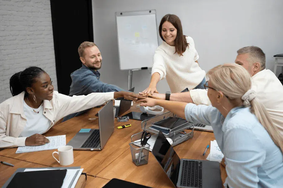 Equipe diversa em reunião no escritório, com participantes ao redor de uma mesa fazendo um gesto de união e colaboração, com laptops e quadro branco ao fundo.