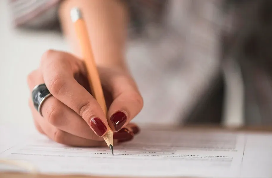 Pessoa escrevendo com lápis sobre uma folha de papel, com close nas mãos e unhas bem cuidadas, em um ambiente de estudo ou trabalho