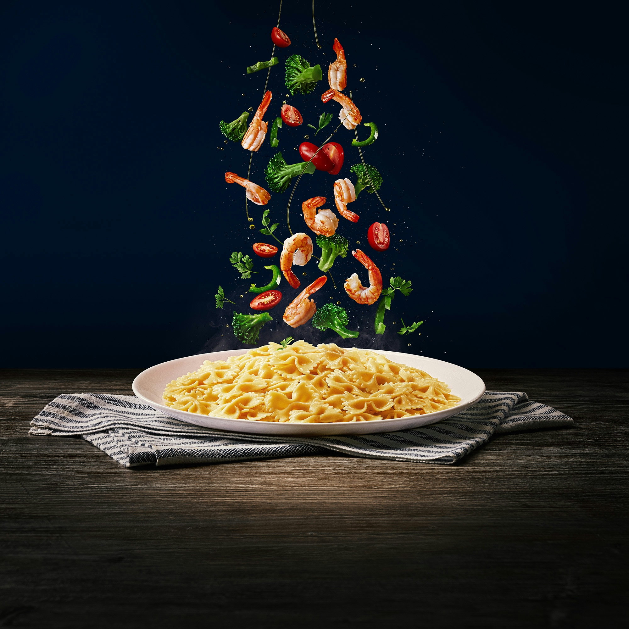Ingredientes (brócolis, tomates cereja, azeite e camarão) caindo em cima do macarrão tipo gravata