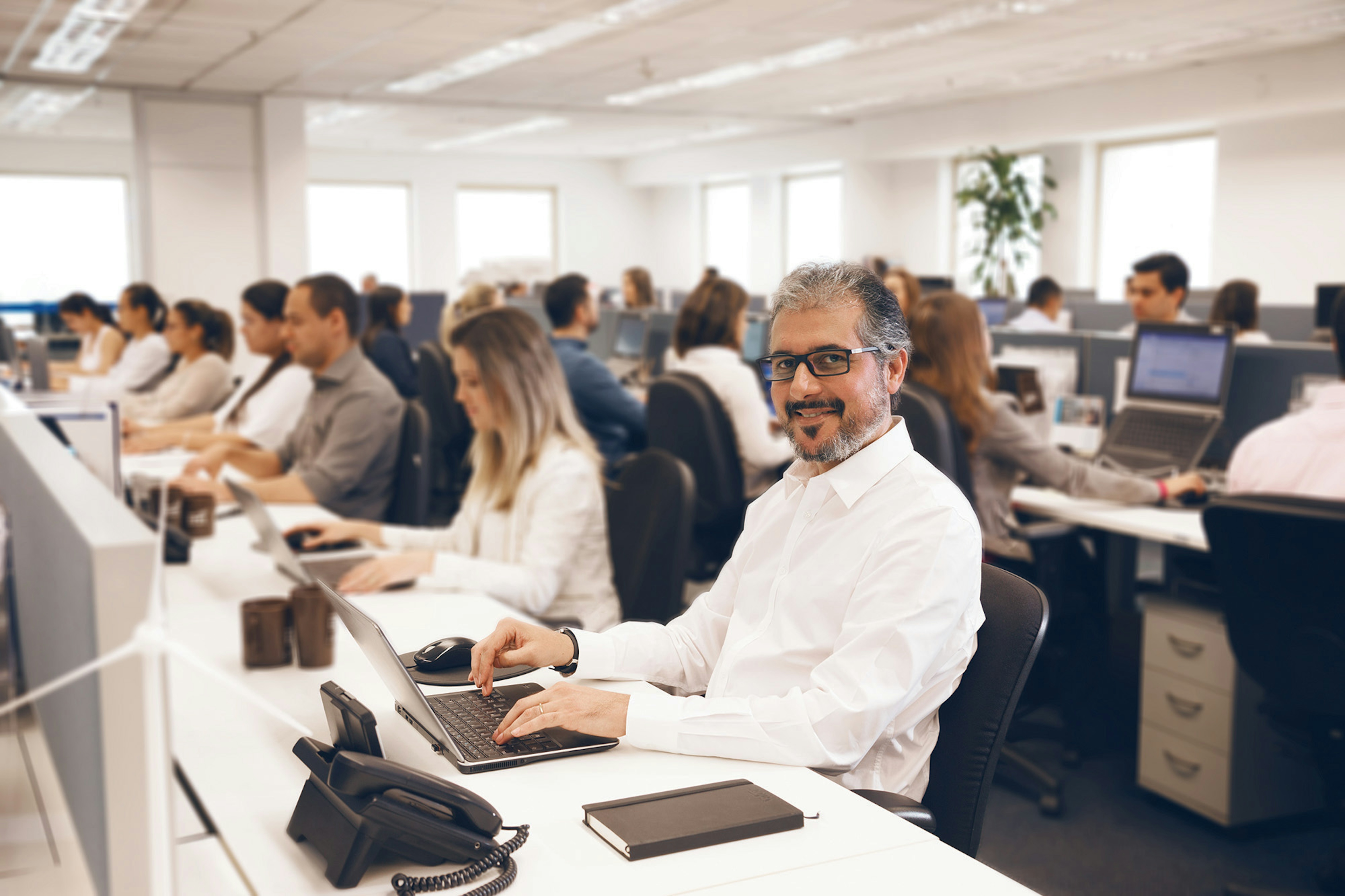 Homem de camisa branca sentado no escritório cheio usando um computador sorrindo para a câmera