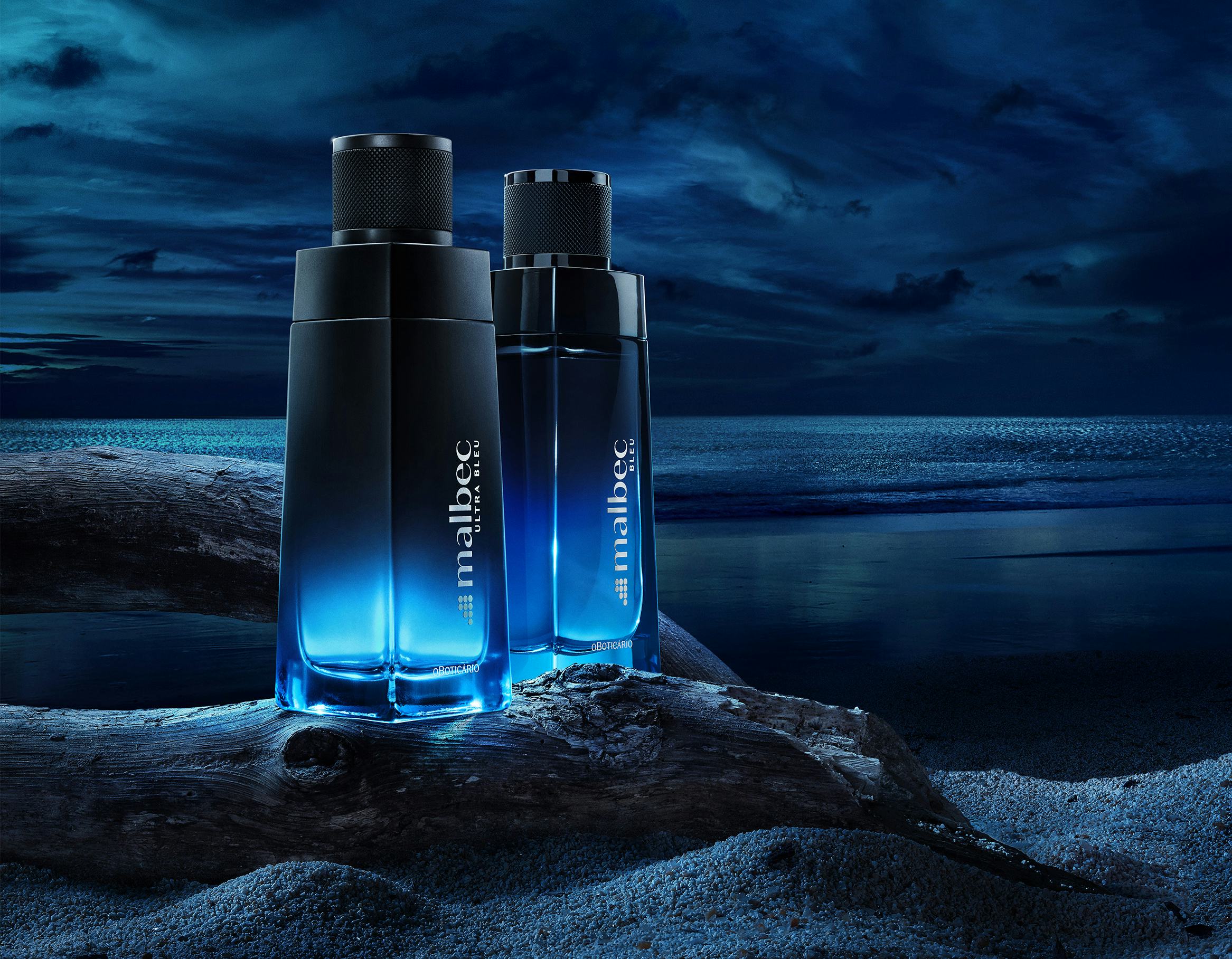 Dois frascos do perfume Malbec Bleu do o Boticário em cima de uma tora de madeira na areia da praia de noite