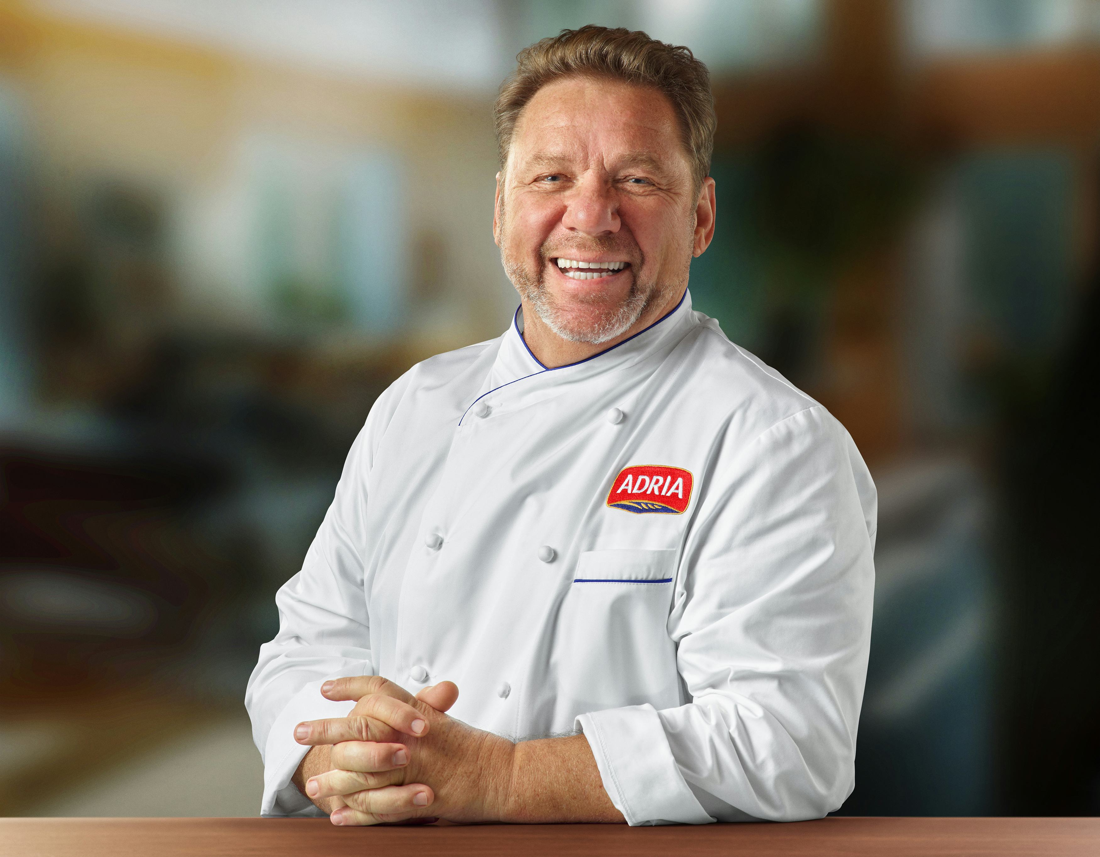 Chefe Claude Troisgros sorrindo com as mãos cruzadas em cima de uma mesa de madeira