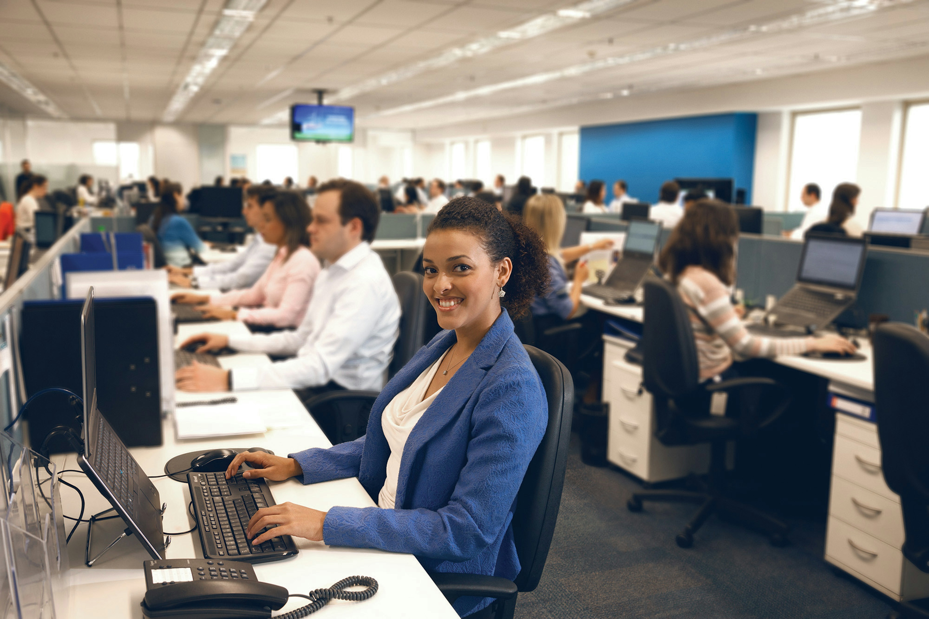 Mulher de blazer azul sentada no escritório cheio usando um computador sorrindo para a câmera