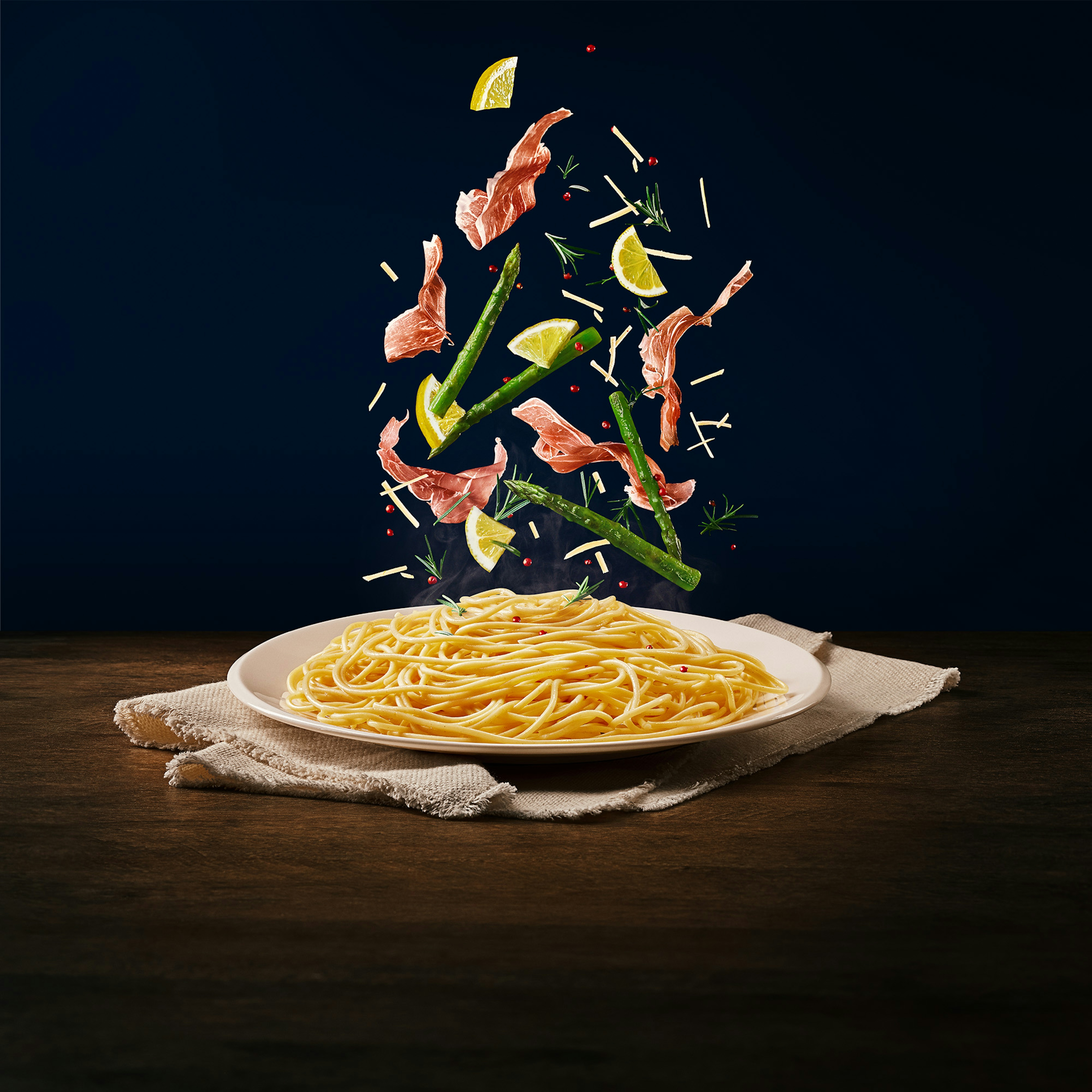Ingredientes (aspargos, limão e presunto parma) caindo em cima do macarrão tipo espaguete