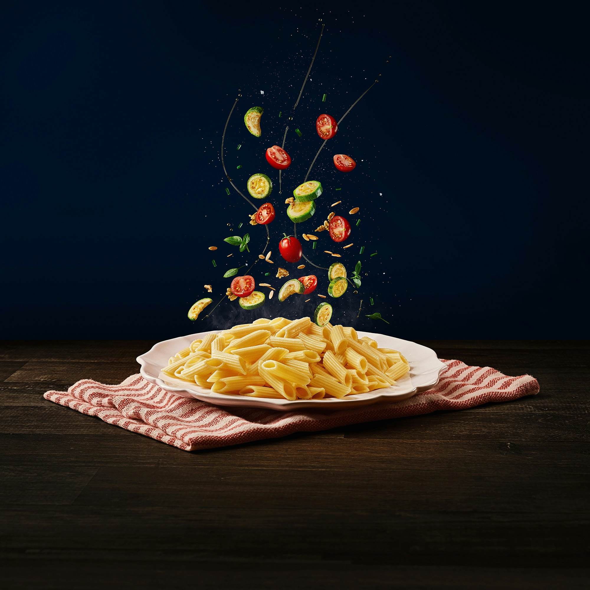Ingredientes (abobrinha, tomates cereja, azeite e grãos) caindo em cima do macarrão tipo penne