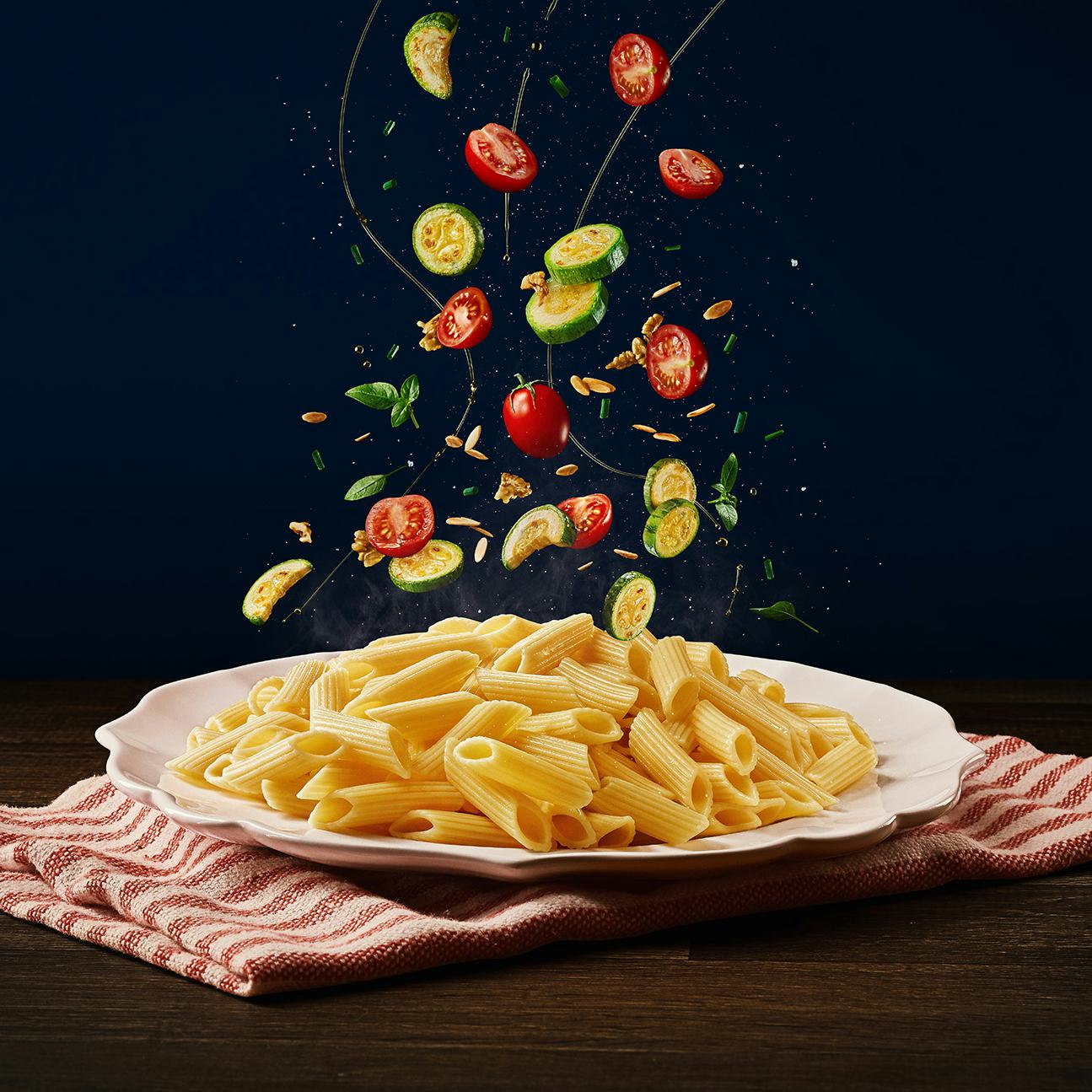 Ingredientes (abobrinha, tomates cereja, azeite e grãos) caindo em cima do macarrão tipo penne