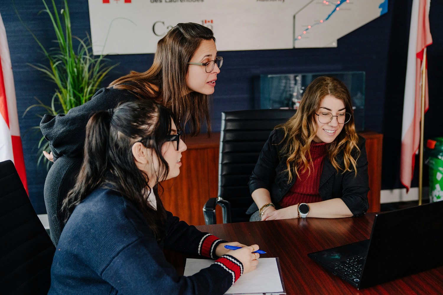 Trois employées du Pilotage St-Laurent collaborent autour d’un ordinateur portable dans une salle de réunion. L’une prend des notes, une autre observe l’écran attentivement, et la troisième sourit, créant une ambiance de travail concentrée et conviviale.