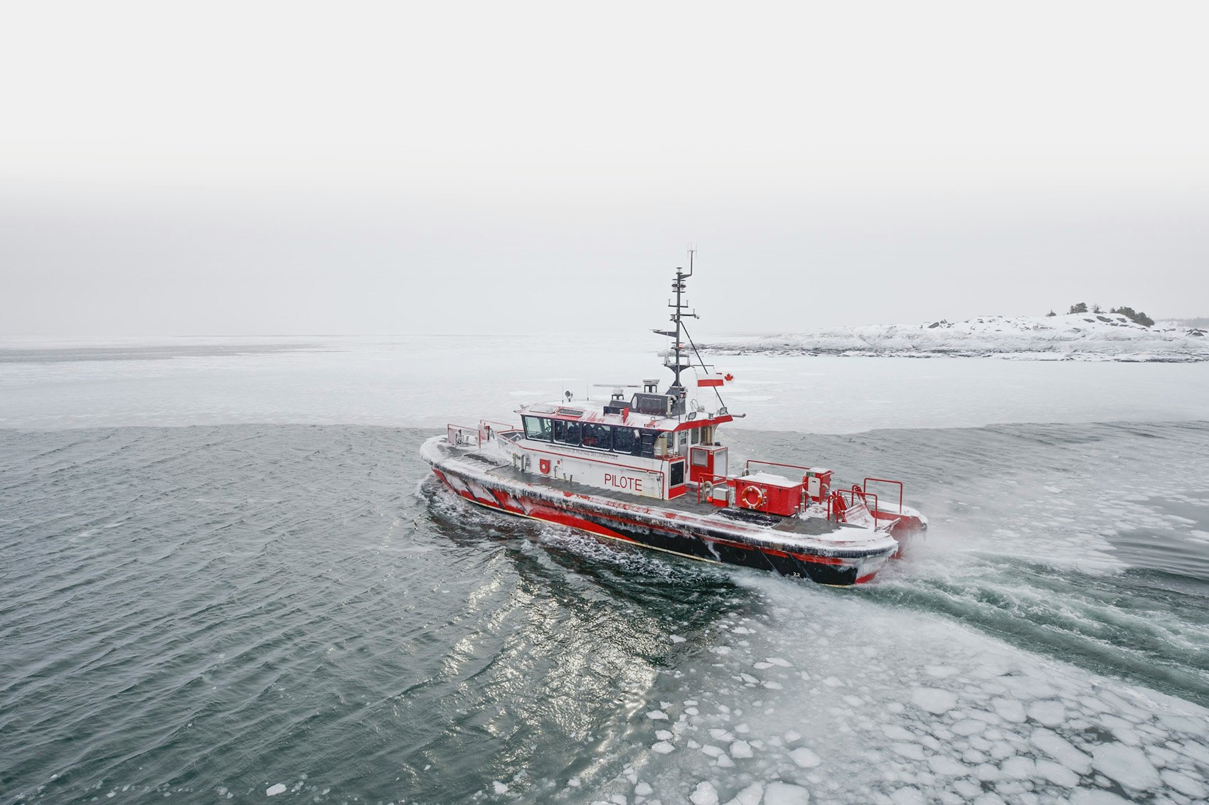 A red-and-white Pilotage St. Lawrence vessel travels through partially frozen waters. The hull and deck are coated in ice, with a snowy shoreline visible in the distance under a grey winter sky.