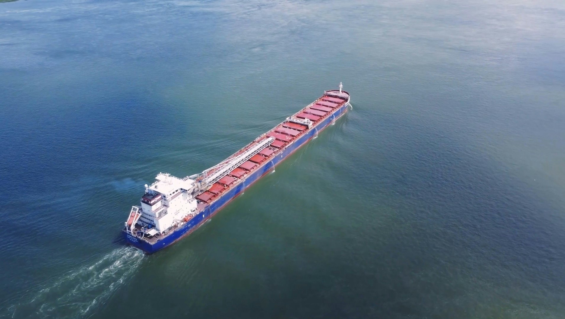 Vue aérienne d’un grand navire de charge bleu et rouge naviguant sur le fleuve Saint-Laurent. Le bateau avance calmement sur une vaste étendue d’eau bleue sous un ciel clair, illustrant la navigation commerciale encadrée par le Pilotage St-Laurent.