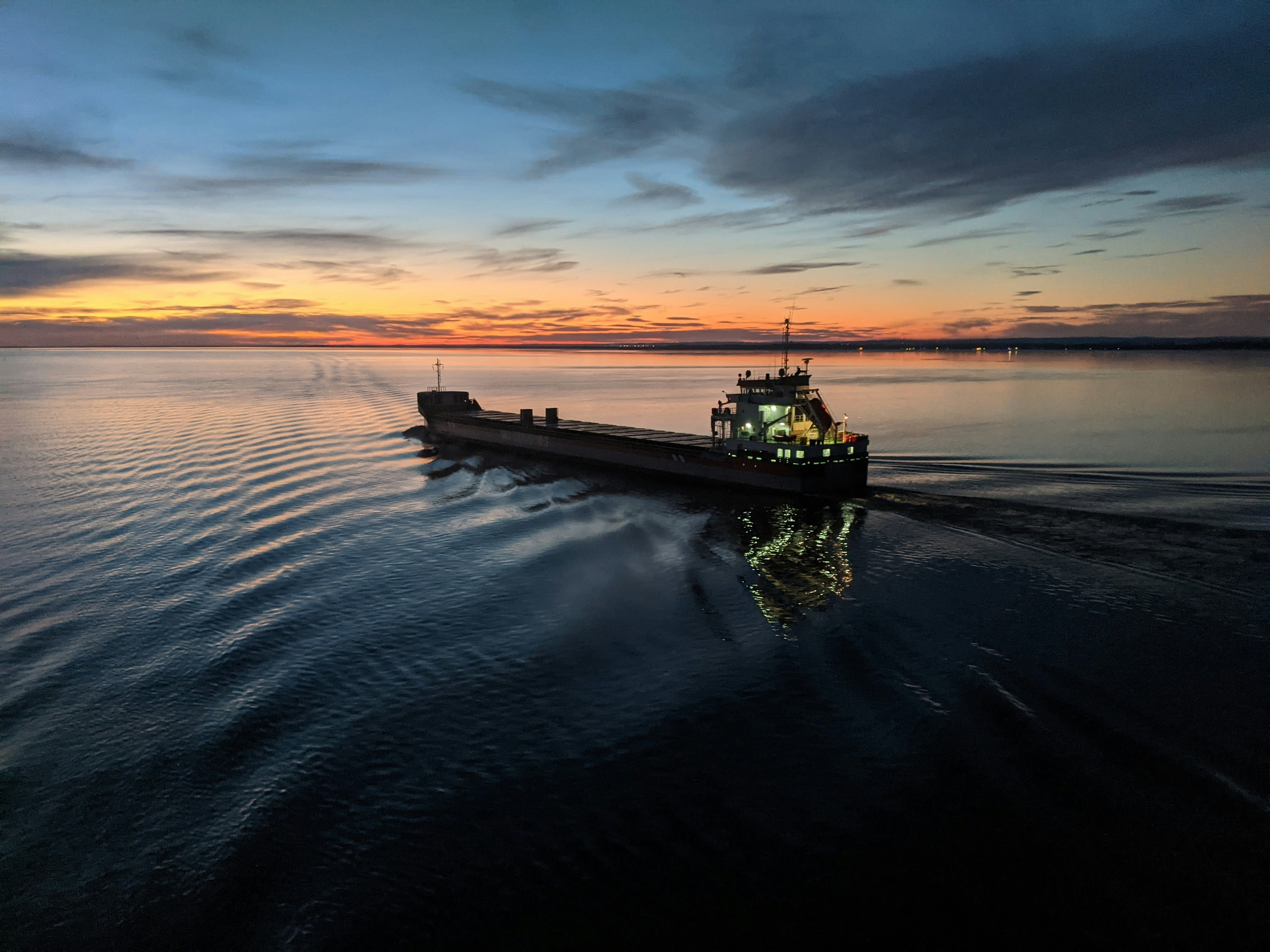 Un grand navire navigue sur le fleuve Saint-Laurent au crépuscule. Les lumières du bateau se reflètent sur l’eau calme tandis que le ciel, aux teintes orangées et bleues, annonce la tombée de la nuit.