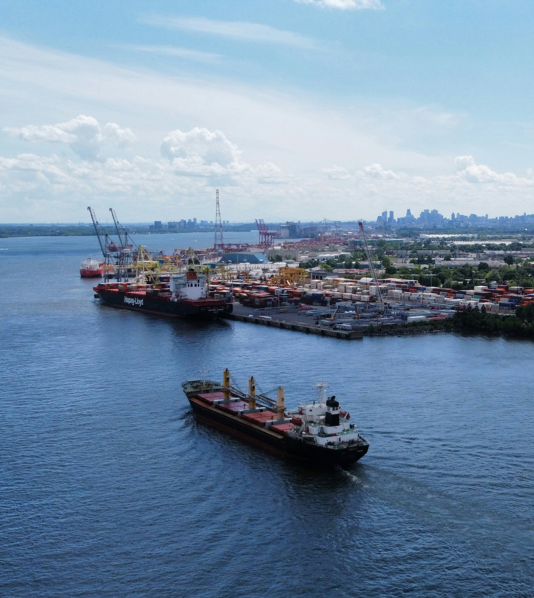 Vue aérienne du port de Montréal. Au premier plan, un grand navire cargo se déplace sur le fleuve Saint-Laurent. À gauche, un autre navire, aux couleurs de Hapag-Lloyd, est amarré au quai près de nombreuses grues portuaires. À l’arrière-plan, on aperçoit une vaste zone de conteneurs ainsi que le centre-ville de Montréal sous un ciel partiellement nuageux.
