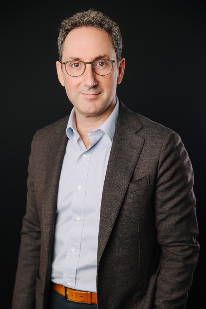 Portrait professionnel d’un homme portant des lunettes, une chemise pâle et un veston foncé, posé devant un fond noir.