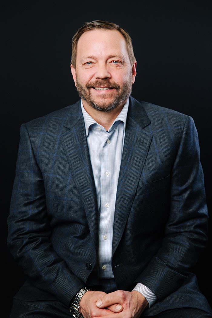 Portrait professionnel d’un homme portant une chemise pâle et un veston foncé, posé devant un fond noir.